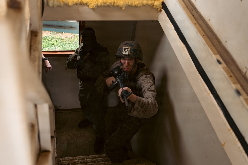 Two military personnel in combat gear are ascending a staircase inside a building, appearing to conduct a room-clearing or building-entry drill. The foreground soldier wears MARPAT (Marine pattern) camouflage and a helmet with a visible insignia patch, holding a rifle in a ready position. The scene appears to be a military training exercise based on the controlled environment.