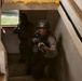 Two military personnel in combat gear are ascending a staircase inside a building, appearing to conduct a room-clearing or building-entry drill. The foreground soldier wears MARPAT (Marine pattern) camouflage and a helmet with a visible insignia patch, holding a rifle in a ready position. The scene appears to be a military training exercise based on the controlled environment.