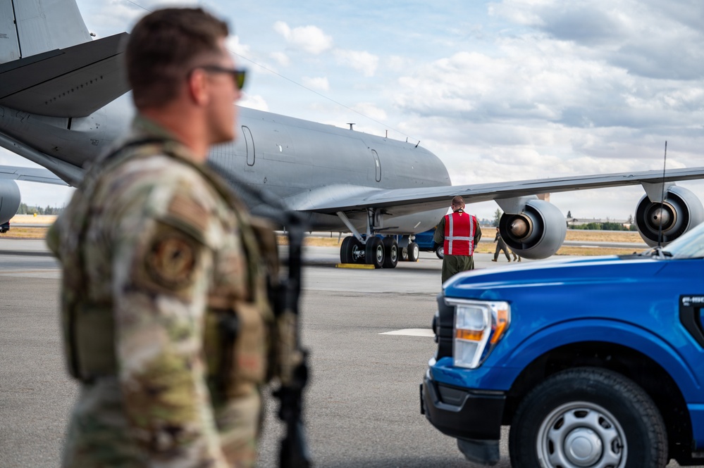 A military flight line scene showing ground operations around a large gray military aircraft, likely a KC-135 or similar tanker/transport. A uniformed service member in multicam/OCP camouflage stands in the foreground while a ground crew member in a red safety vest marshals or inspects the aircraft. A blue Ford F-150 pickup truck is parked nearby on the tarmac.