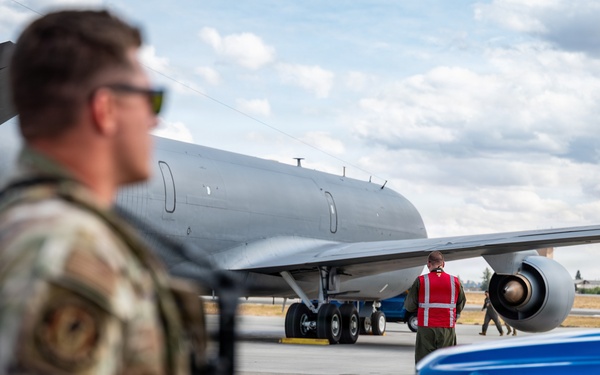 A military flight line scene showing ground operations around a large gray military aircraft, likely a KC-135 or similar tanker/transport. A uniformed service member in multicam/OCP camouflage stands in the foreground while a ground crew member in a red safety vest marshals or inspects the aircraft. A blue Ford F-150 pickup truck is parked nearby on the tarmac.