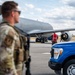 A military flight line scene showing ground operations around a large gray military aircraft, likely a KC-135 or similar tanker/transport. A uniformed service member in multicam/OCP camouflage stands in the foreground while a ground crew member in a red safety vest marshals or inspects the aircraft. A blue Ford F-150 pickup truck is parked nearby on the tarmac.