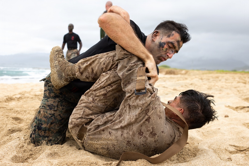 Two military personnel in MARPAT desert camouflage uniforms are engaged in close-quarters combat training on a sandy beach. One individual wearing camouflage face paint has the other in a ground grappling position, consistent with Marine Corps Martial Arts Program (MCMAP) training. A third uniformed individual observing in the background suggests this is a supervised training exercise.