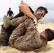 Two military personnel in MARPAT desert camouflage uniforms are engaged in close-quarters combat training on a sandy beach. One individual wearing camouflage face paint has the other in a ground grappling position, consistent with Marine Corps Martial Arts Program (MCMAP) training. A third uniformed individual observing in the background suggests this is a supervised training exercise.