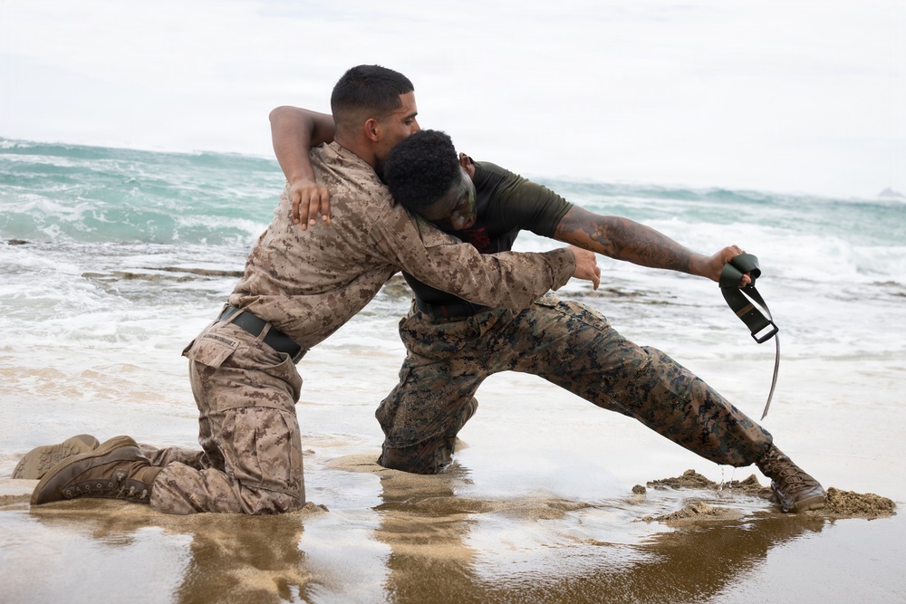 Two military personnel in U.S. Marine Corps camouflage uniforms are engaged in a close-quarters combat training exercise on a beach shoreline. One Marine is kneeling in shallow water while the other stands in a grappling position, with one individual holding what appears to be a training knife or blade. The setting appears to be a military training environment given the controlled nature of the engagement.