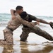 Two military personnel in U.S. Marine Corps camouflage uniforms are engaged in a close-quarters combat training exercise on a beach shoreline. One Marine is kneeling in shallow water while the other stands in a grappling position, with one individual holding what appears to be a training knife or blade. The setting appears to be a military training environment given the controlled nature of the engagement.