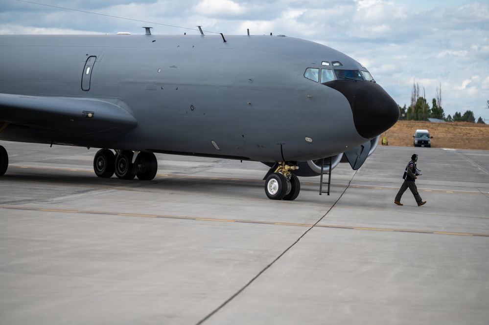 A large military aircraft, likely a KC-135 or similar tanker/reconnaissance variant, is parked on a concrete flight line apron. A single ground crew member is walking near the nose of the aircraft, possibly conducting a pre-flight or post-flight inspection. The aircraft is painted in a dark gray military finish with a black radome on the nose.