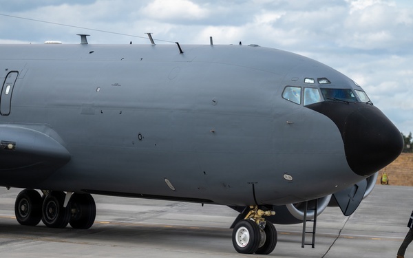 A large military aircraft, likely a KC-135 or similar tanker/reconnaissance variant, is parked on a concrete flight line apron. A single ground crew member is walking near the nose of the aircraft, possibly conducting a pre-flight or post-flight inspection. The aircraft is painted in a dark gray military finish with a black radome on the nose.
