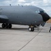 A large military aircraft, likely a KC-135 or similar tanker/reconnaissance variant, is parked on a concrete flight line apron. A single ground crew member is walking near the nose of the aircraft, possibly conducting a pre-flight or post-flight inspection. The aircraft is painted in a dark gray military finish with a black radome on the nose.