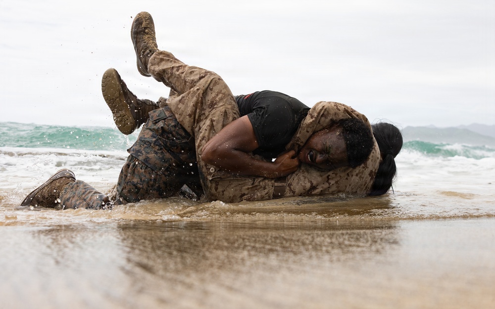 Two individuals in military camouflage uniforms are engaged in close-quarters combat training on a beach shoreline. They are grappling on the wet sand at the water's edge, with waves visible in the background. Both are covered in mud and water, indicating an intensive physical training exercise.