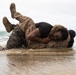 Two individuals in military camouflage uniforms are engaged in close-quarters combat training on a beach shoreline. They are grappling on the wet sand at the water's edge, with waves visible in the background. Both are covered in mud and water, indicating an intensive physical training exercise.