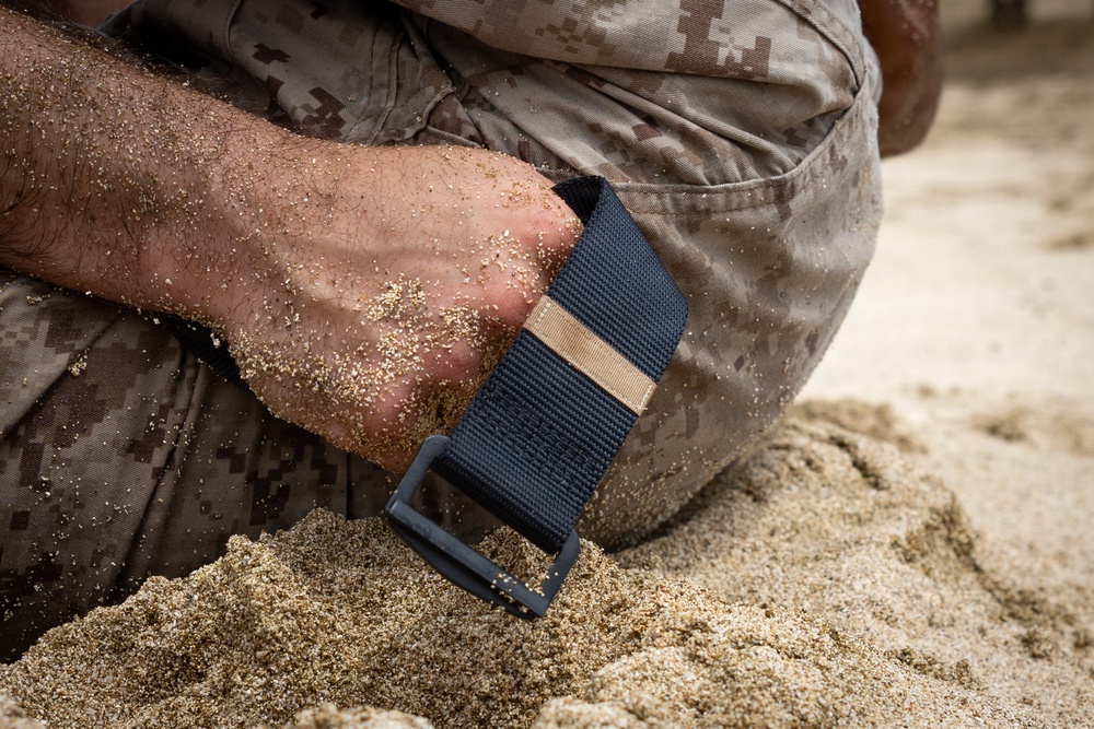 A close-up image showing a person wearing digital camouflage military-pattern clothing kneeling or crouching in sand. Their hand and wrist are heavily coated with sand, and they are wearing a navy blue nylon watch strap or wristband with a dark buckle. The scene suggests a beach or sandy training environment.