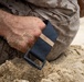 A close-up image showing a person wearing digital camouflage military-pattern clothing kneeling or crouching in sand. Their hand and wrist are heavily coated with sand, and they are wearing a navy blue nylon watch strap or wristband with a dark buckle. The scene suggests a beach or sandy training environment.