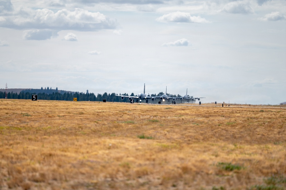 Multiple large military transport aircraft are lined up on a runway, appearing to be taxiing or preparing for takeoff in a formation departure. The scene is set at a military airfield surrounded by dry grassland and a treeline in the background. Runway distance markers (numbered 3, 4, 5) are visible along the taxiway edge.