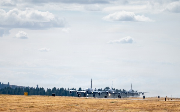 Multiple large military transport aircraft are lined up on a runway, appearing to be taxiing or preparing for takeoff in a formation departure. The scene is set at a military airfield surrounded by dry grassland and a treeline in the background. Runway distance markers (numbered 3, 4, 5) are visible along the taxiway edge.