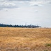 Multiple large military transport aircraft are lined up on a runway, appearing to be taxiing or preparing for takeoff in a formation departure. The scene is set at a military airfield surrounded by dry grassland and a treeline in the background. Runway distance markers (numbered 3, 4, 5) are visible along the taxiway edge.