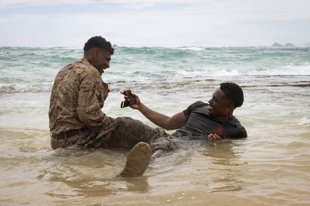 Two individuals are sitting in shallow ocean surf near a beach shoreline. One person is wearing a military camouflage uniform with combat boots, while the other is in civilian dark clothing. They appear to be interacting and sharing a moment, with one handing or showing a small device (possibly a phone) to the other.