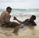 Two individuals are sitting in shallow ocean surf near a beach shoreline. One person is wearing a military camouflage uniform with combat boots, while the other is in civilian dark clothing. They appear to be interacting and sharing a moment, with one handing or showing a small device (possibly a phone) to the other.