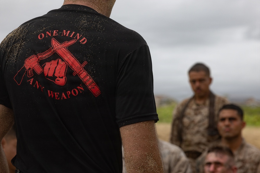 A person wearing a black t-shirt with a red graphic reading 'ONE MIND ANY WEAPON' stands with their back to the camera, covered in sand or dirt. In the background, several individuals in military camouflage uniforms are visible, slightly out of focus. The setting appears to be an outdoor training environment under an overcast sky.