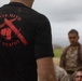 A person wearing a black t-shirt with a red graphic reading 'ONE MIND ANY WEAPON' stands with their back to the camera, covered in sand or dirt. In the background, several individuals in military camouflage uniforms are visible, slightly out of focus. The setting appears to be an outdoor training environment under an overcast sky.