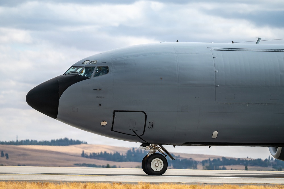 A large military aircraft, consistent with a KC-135 Stratotanker or similar tanker/transport aircraft, is taxiing on a runway. The aircraft is painted in a flat gray military finish with a black radome nose cone. The setting appears to be a military airfield surrounded by rolling hills and sparse tree lines under a partly cloudy sky.