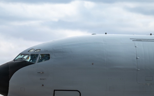 A large military aircraft, consistent with a KC-135 Stratotanker or similar tanker/transport aircraft, is taxiing on a runway. The aircraft is painted in a flat gray military finish with a black radome nose cone. The setting appears to be a military airfield surrounded by rolling hills and sparse tree lines under a partly cloudy sky.