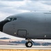 A large military aircraft, consistent with a KC-135 Stratotanker or similar tanker/transport aircraft, is taxiing on a runway. The aircraft is painted in a flat gray military finish with a black radome nose cone. The setting appears to be a military airfield surrounded by rolling hills and sparse tree lines under a partly cloudy sky.