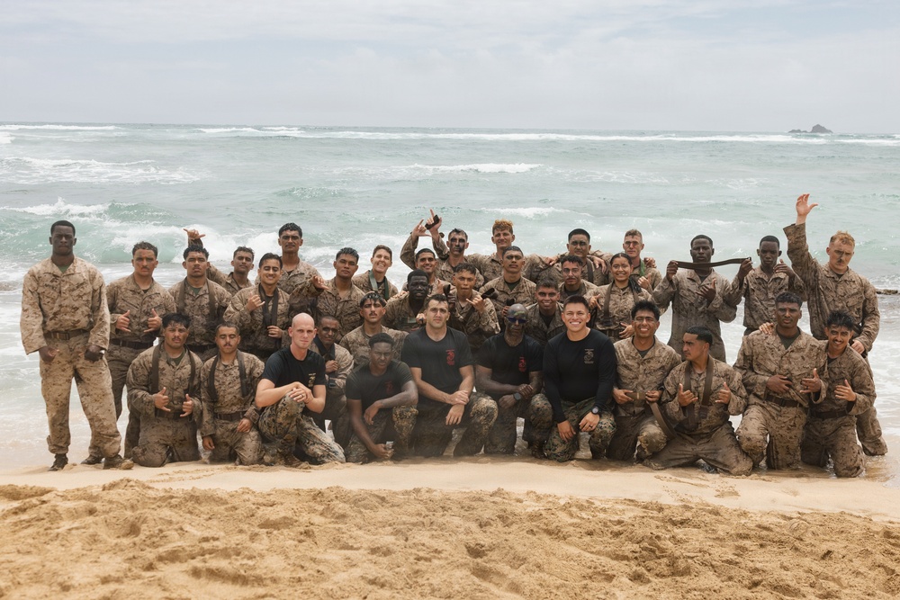 A large group of military personnel, appearing to be U.S. Marines based on their desert camouflage uniforms, pose for a group photo on a sandy beach with ocean waves visible in the background. The group includes both uniformed members and a few individuals in black t-shirts who appear to be instructors or leaders. The personnel appear wet and sandy, suggesting they recently completed a water-based training exercise.