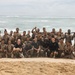 A large group of military personnel, appearing to be U.S. Marines based on their desert camouflage uniforms, pose for a group photo on a sandy beach with ocean waves visible in the background. The group includes both uniformed members and a few individuals in black t-shirts who appear to be instructors or leaders. The personnel appear wet and sandy, suggesting they recently completed a water-based training exercise.
