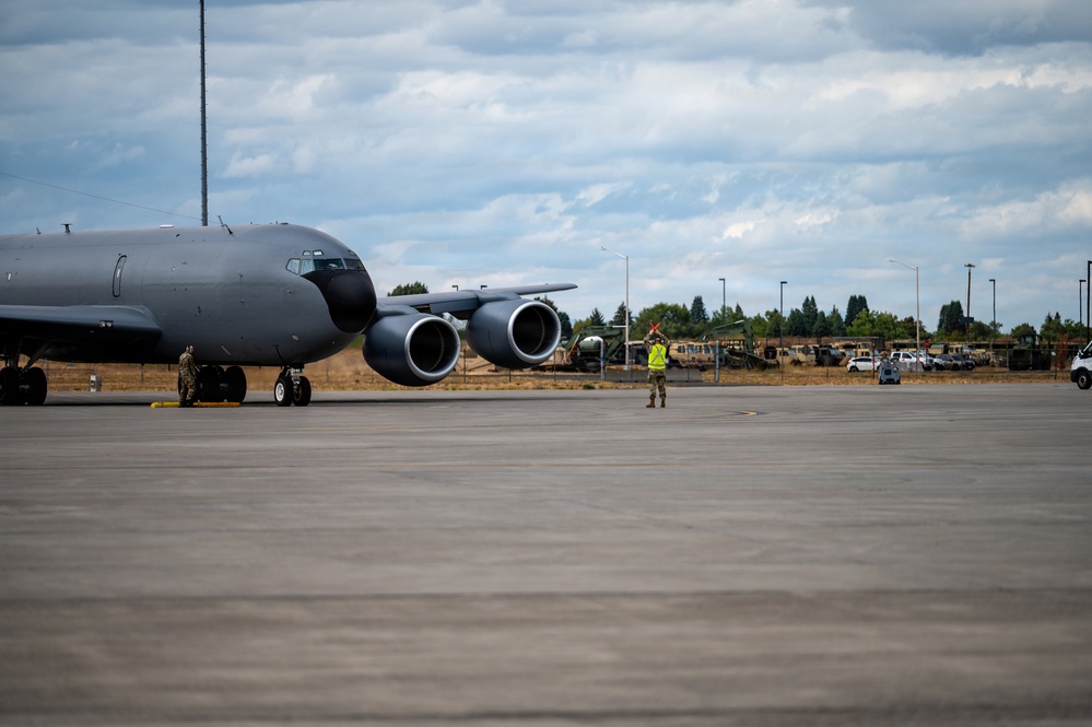 A large military aircraft, consistent with a KC-135 or similar tanker/transport, is parked on a concrete flight line at a military airfield. A ground crew member wearing a high-visibility yellow vest is marshaling the aircraft using signal wands. The scene appears to be a routine ground operations activity at what looks like a U.S. Air Force installation.