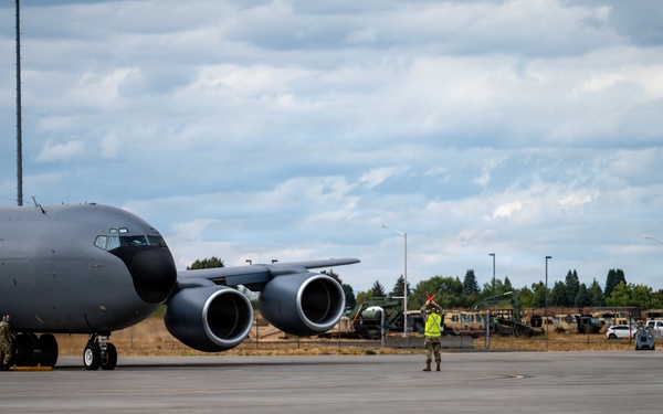 A large military aircraft, consistent with a KC-135 or similar tanker/transport, is parked on a concrete flight line at a military airfield. A ground crew member wearing a high-visibility yellow vest is marshaling the aircraft using signal wands. The scene appears to be a routine ground operations activity at what looks like a U.S. Air Force installation.