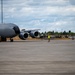 A large military aircraft, consistent with a KC-135 or similar tanker/transport, is parked on a concrete flight line at a military airfield. A ground crew member wearing a high-visibility yellow vest is marshaling the aircraft using signal wands. The scene appears to be a routine ground operations activity at what looks like a U.S. Air Force installation.