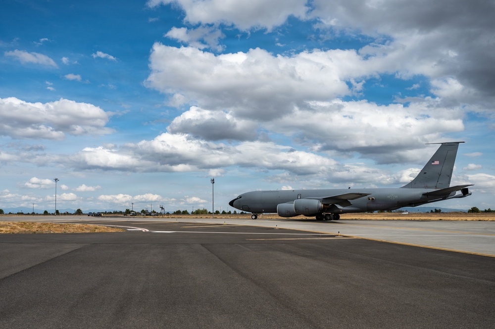 A large gray military aircraft, consistent with a KC-135 Stratotanker, is parked or taxiing on a tarmac at a military airfield. An American flag is visible on the tail fin, indicating U.S. Air Force ownership. The setting appears to be a dry, arid region under a partly cloudy blue sky.