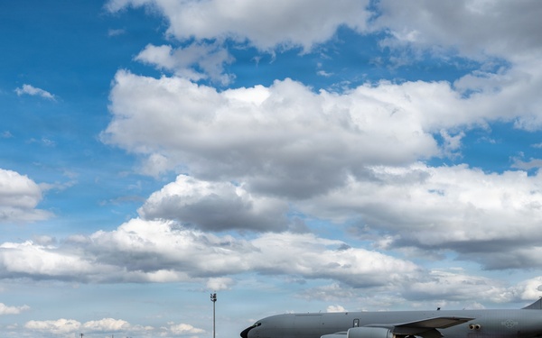 A large gray military aircraft, consistent with a KC-135 Stratotanker, is parked or taxiing on a tarmac at a military airfield. An American flag is visible on the tail fin, indicating U.S. Air Force ownership. The setting appears to be a dry, arid region under a partly cloudy blue sky.