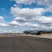 A large gray military aircraft, consistent with a KC-135 Stratotanker, is parked or taxiing on a tarmac at a military airfield. An American flag is visible on the tail fin, indicating U.S. Air Force ownership. The setting appears to be a dry, arid region under a partly cloudy blue sky.
