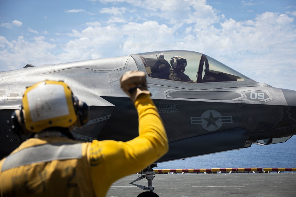 A deck crew member in yellow flight deck gear signals to a pilot seated in an F-35B Lightning II stealth fighter jet, numbered '09', on what appears to be the flight deck of a naval aircraft carrier. The US military roundel insignia is clearly visible on the fuselage. The scene takes place under partly cloudy skies with open ocean visible in the background.