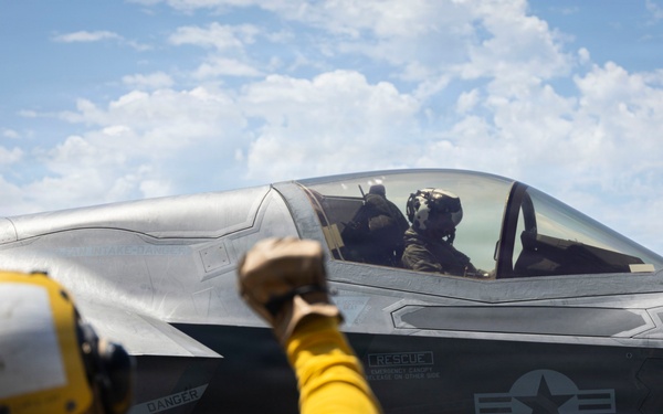 A deck crew member in yellow flight deck gear signals to a pilot seated in an F-35B Lightning II stealth fighter jet, numbered '09', on what appears to be the flight deck of a naval aircraft carrier. The US military roundel insignia is clearly visible on the fuselage. The scene takes place under partly cloudy skies with open ocean visible in the background.