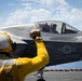 A deck crew member in yellow flight deck gear signals to a pilot seated in an F-35B Lightning II stealth fighter jet, numbered '09', on what appears to be the flight deck of a naval aircraft carrier. The US military roundel insignia is clearly visible on the fuselage. The scene takes place under partly cloudy skies with open ocean visible in the background.