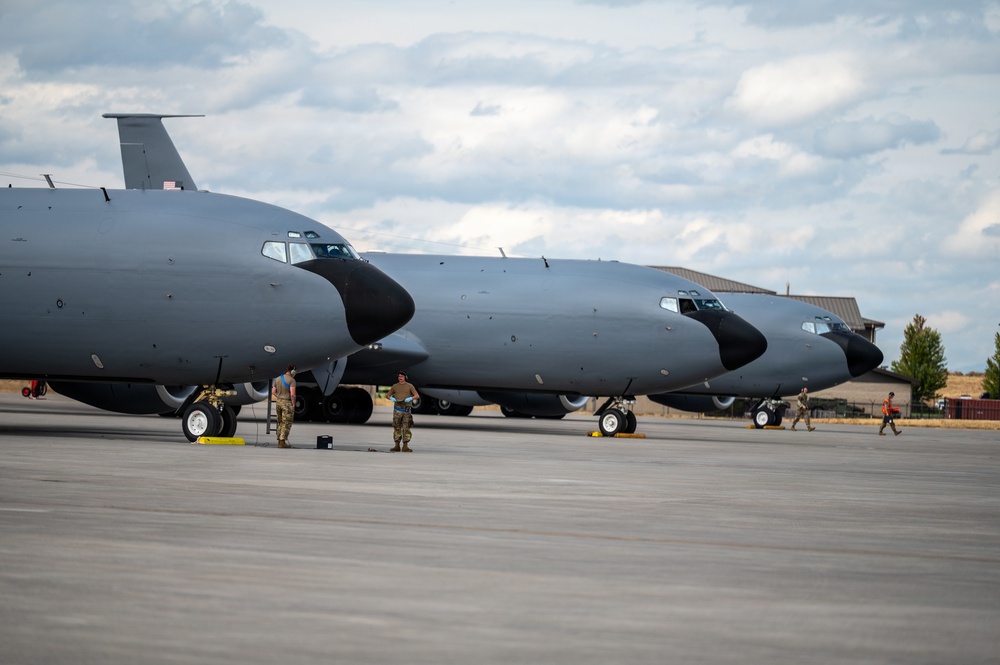 Multiple large military aircraft, consistent with KC-135 Stratotanker aerial refueling tankers, are parked on a concrete flight line apron. Ground crew personnel in camouflage uniforms are conducting pre-flight or maintenance checks around the aircraft. The setting appears to be a U.S. Air Force base with hangars and trees visible in the background.