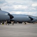Multiple large military aircraft, consistent with KC-135 Stratotanker aerial refueling tankers, are parked on a concrete flight line apron. Ground crew personnel in camouflage uniforms are conducting pre-flight or maintenance checks around the aircraft. The setting appears to be a U.S. Air Force base with hangars and trees visible in the background.