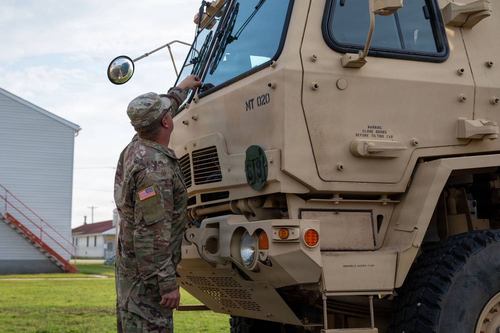A U.S. Army soldier in MultiCam/OCP camouflage uniform is conducting a pre-operation inspection of a large tan military tactical vehicle, identified as 'MT 020'. The vehicle appears to be a Heavy Expanded Mobility Tactical Truck (HEMTT) or similar large military transport vehicle with desert tan paint. The scene is set at what appears to be a military installation with buildings visible in the background.