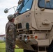 A U.S. Army soldier in MultiCam/OCP camouflage uniform is conducting a pre-operation inspection of a large tan military tactical vehicle, identified as 'MT 020'. The vehicle appears to be a Heavy Expanded Mobility Tactical Truck (HEMTT) or similar large military transport vehicle with desert tan paint. The scene is set at what appears to be a military installation with buildings visible in the background.