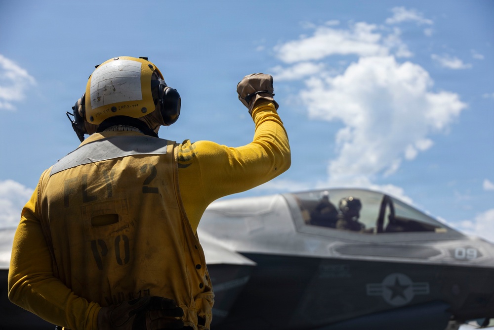 A flight deck crew member in yellow gear and a flight helmet gives a raised fist signal to an F-35 fighter jet on what appears to be an aircraft carrier deck. The aircraft bears U.S. military markings and the number '09' on its fuselage. A pilot is visible in the cockpit of the aircraft.