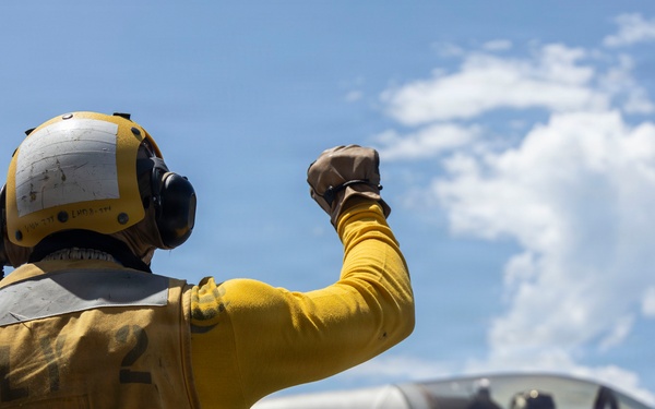 A flight deck crew member in yellow gear and a flight helmet gives a raised fist signal to an F-35 fighter jet on what appears to be an aircraft carrier deck. The aircraft bears U.S. military markings and the number '09' on its fuselage. A pilot is visible in the cockpit of the aircraft.