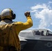 A flight deck crew member in yellow gear and a flight helmet gives a raised fist signal to an F-35 fighter jet on what appears to be an aircraft carrier deck. The aircraft bears U.S. military markings and the number '09' on its fuselage. A pilot is visible in the cockpit of the aircraft.