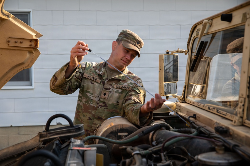 A U.S. Army soldier named Wygle is performing vehicle maintenance on a military HMMWV (Humvee), checking the oil dipstick on the open engine compartment. The soldier is wearing MultiCam (OCP) camouflage uniform with clearly visible name tape 'WYGLE' and 'U.S. ARMY' tape. The image is taken outdoors near a white building in good lighting conditions.