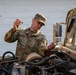 A U.S. Army soldier named Wygle is performing vehicle maintenance on a military HMMWV (Humvee), checking the oil dipstick on the open engine compartment. The soldier is wearing MultiCam (OCP) camouflage uniform with clearly visible name tape 'WYGLE' and 'U.S. ARMY' tape. The image is taken outdoors near a white building in good lighting conditions.