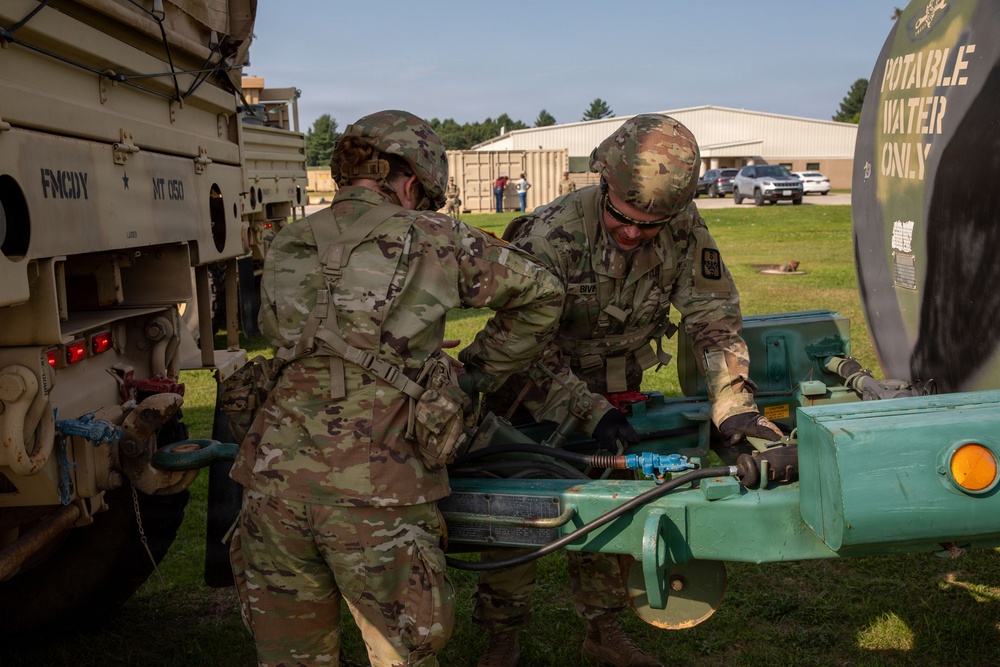 Two soldiers in OCP (Operational Camouflage Pattern) uniforms are working together on a potable water trailer, connecting or adjusting hoses and fittings. The scene appears to be a military training or operational area with military vehicles and a building visible in the background. The activity suggests water distribution or equipment setup operations.