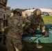 Two soldiers in OCP (Operational Camouflage Pattern) uniforms are working together on a potable water trailer, connecting or adjusting hoses and fittings. The scene appears to be a military training or operational area with military vehicles and a building visible in the background. The activity suggests water distribution or equipment setup operations.