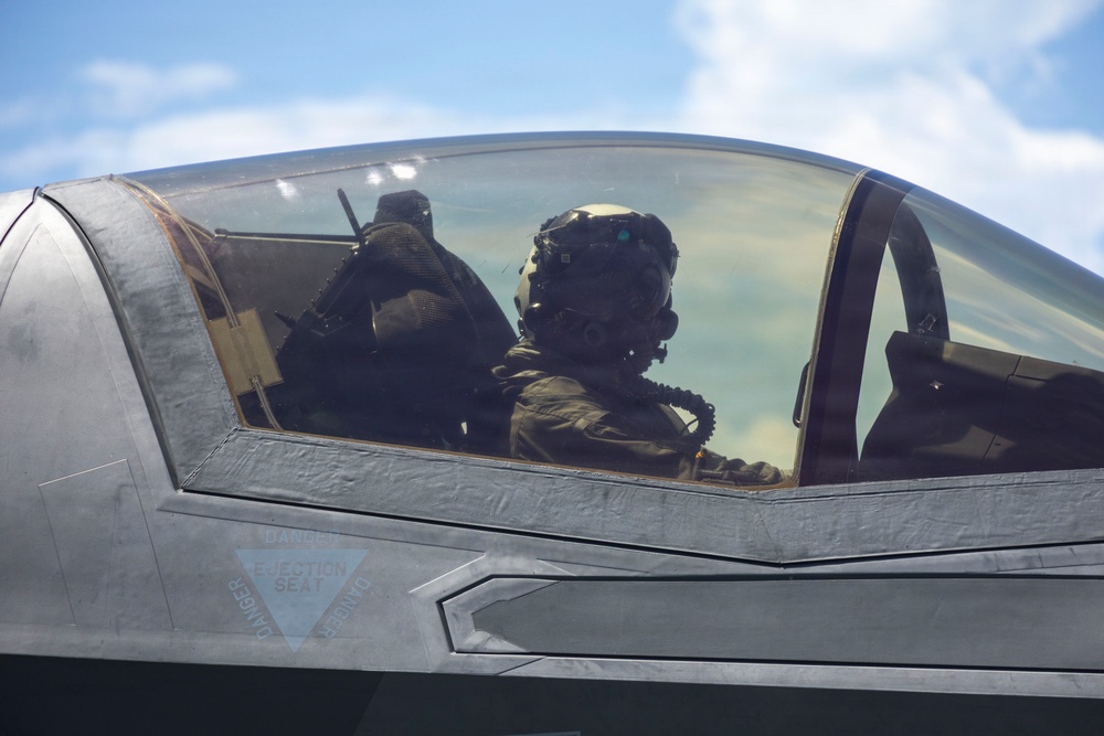 A military pilot is seated in the cockpit of a modern stealth fighter aircraft, identifiable by its low-observable canopy design and warning markings. The pilot is wearing a full flight helmet with oxygen mask and flight suit. The image is taken from outside the closed canopy, looking in at an angle against a partly cloudy sky.