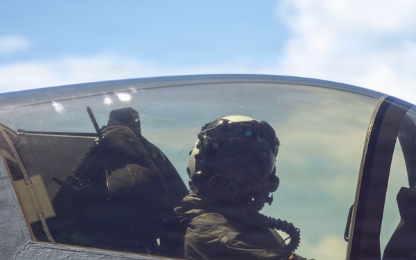 A military pilot is seated in the cockpit of a modern stealth fighter aircraft, identifiable by its low-observable canopy design and warning markings. The pilot is wearing a full flight helmet with oxygen mask and flight suit. The image is taken from outside the closed canopy, looking in at an angle against a partly cloudy sky.